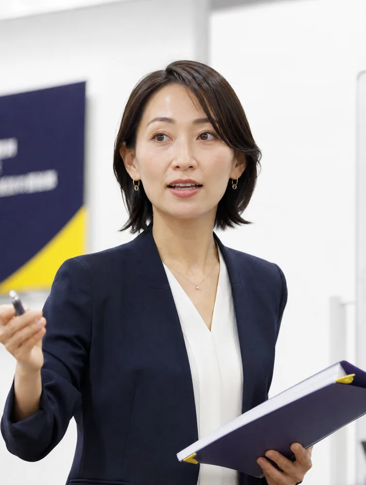 デモ用の生成画像（架空のファシリテーターの写真）。ジャケット姿の女性が資料を手に、参加者の方へ視線を向けている。